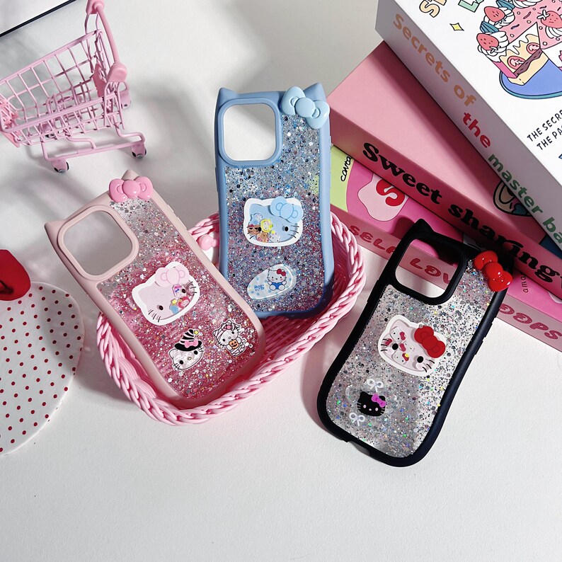 Może przedstawiać: Trzy etui na telefon z motywami Hello Kitty i brokatem. R&oacute;żowe, niebieskie i czarne etui są prezentowane w r&oacute;żowym koszyku. R&oacute;żowy w&oacute;zek na zakupy jest widoczny w tle.