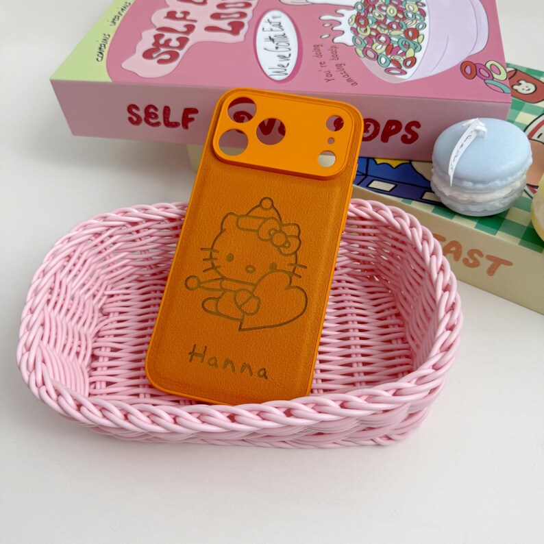 May include: An orange phone case with a Hello Kitty design and the name "Hanna" in a pink woven basket. The case has an orange camera lens protector. A box with the word "SELF" is in the background.