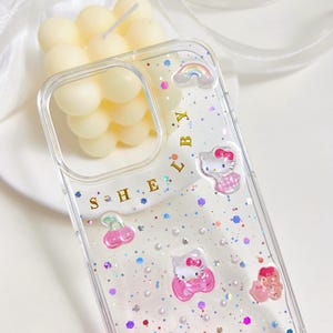 May include: Clear phone case with glitter and Hello Kitty designs. The case features a rainbow, cherries, and the text "SHE LBY". The case is designed to fit a phone and has a clear, transparent appearance.