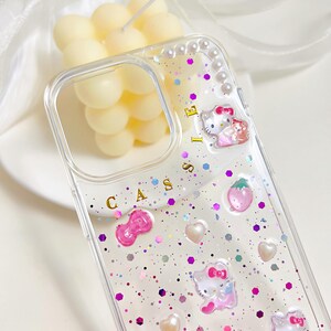 May include: Clear phone case with glitter, pink accents, and a cartoon cat design. The case features a bow, hearts, and a strawberry. The word "CASSIE" is printed on the case.