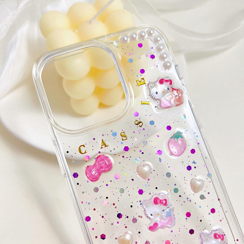 May include: Clear phone case with glitter, pink accents, and a cartoon cat design. The case features a bow, hearts, and a strawberry. The word "CASSIE" is printed on the case.