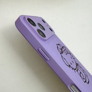 Puede incluir: Una funda de tel&eacute;fono lavanda con un contorno negro de un personaje de dibujos animados. La funda tiene un recorte para la c&aacute;mara y detalles de botones. La funda est&aacute; hecha de un material liso.