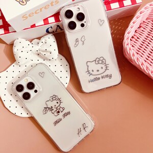 May include: Two clear phone cases with Hello Kitty designs. One case features a Hello Kitty graphic with a bow and the words "Hello Kitty." The other case has a Hello Kitty graphic and the initials "B&G." A white bow with black polka dots and a pink basket are also in the image.