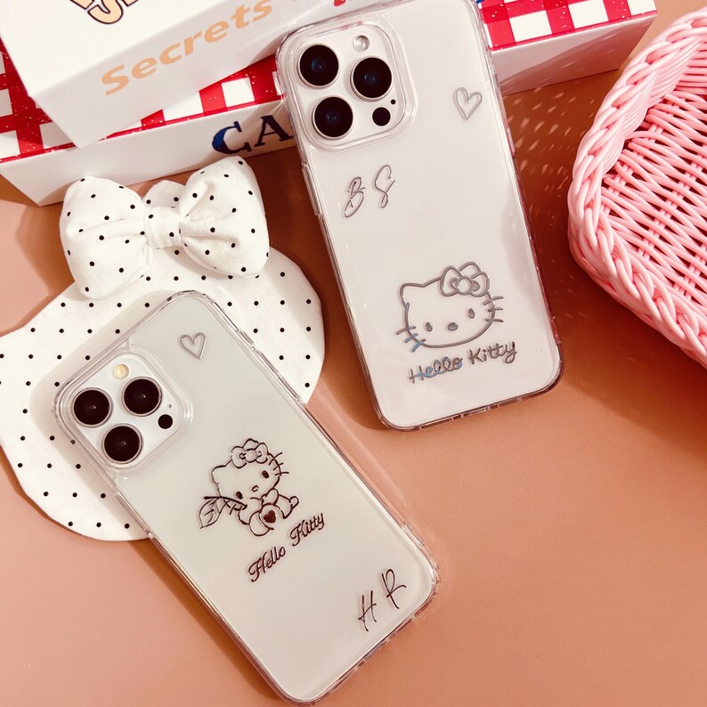 May include: Two clear phone cases with Hello Kitty designs. One case features a Hello Kitty graphic with a bow and the words "Hello Kitty." The other case has a Hello Kitty graphic and the initials "B&G." A white bow with black polka dots and a pink basket are also in the image.
