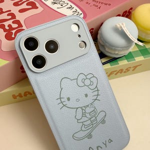 Puede incluir: Funda de tel&eacute;fono azul claro con un dise&ntilde;o de Hello Kitty en monopat&iacute;n. La funda tiene un m&oacute;dulo de c&aacute;mara gris y el nombre "Anya" est&aacute; impreso en la parte inferior. La funda est&aacute; sobre una caja rosa y amarilla.