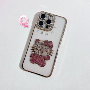 K&ouml;nnte beinhalten: Wei&szlig;es iPhone-Case mit goldfarbenem Rand und einem Hello-Kitty-Design aus Strasssteinen. Das Case hat eine transparente R&uuml;ckseite und eine Kameraobjektivabdeckung aus Strasssteinen.