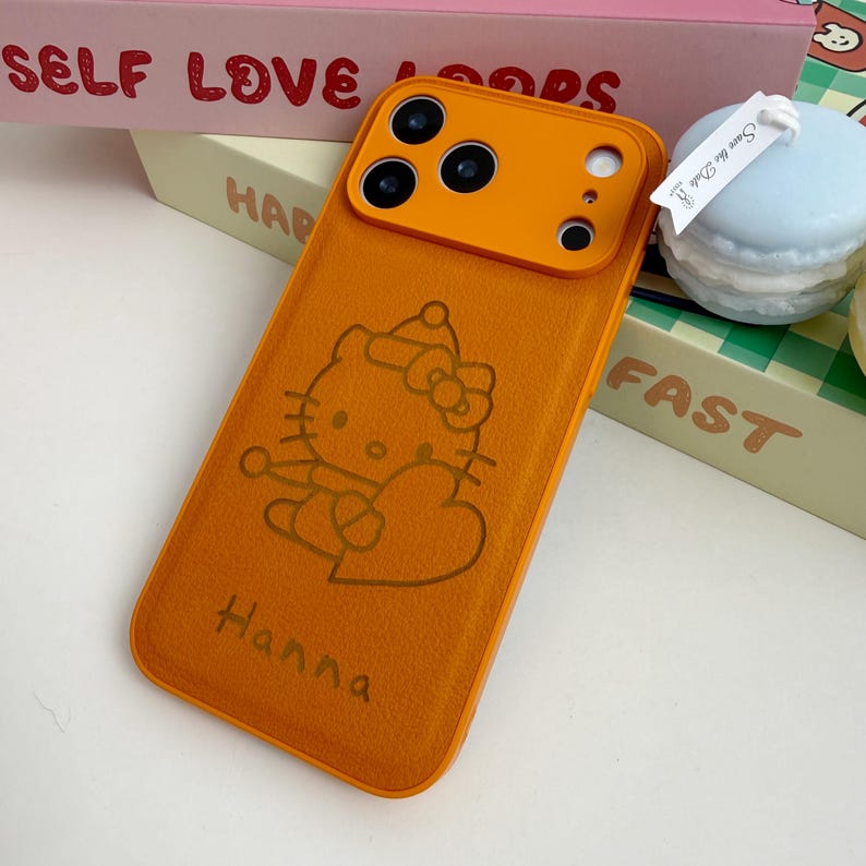 May include: An orange phone case featuring a Hello Kitty design and the name "Hanna". The case has an orange camera module. The background includes the words "SELF LOVE" and "FAST".