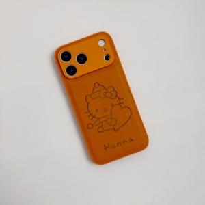 Puede incluir: Funda de tel&eacute;fono naranja con un dise&ntilde;o de Hello Kitty sosteniendo un coraz&oacute;n. La funda tiene un recorte para la c&aacute;mara y el nombre "Hanna" est&aacute; impreso en la parte inferior. La funda est&aacute; hecha de un material texturizado.