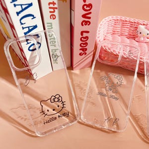 May include: Clear phone cases featuring Hello Kitty designs. One case has a Hello Kitty face, the other has a Hello Kitty angel design. Both cases have the words "Hello Kitty" printed on them. The cases are displayed in front of books and a pink basket.