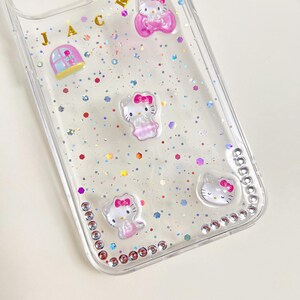 May include: Clear phone case decorated with Hello Kitty figures, pink bows, and glitter. The case has a border of small, clear rhinestones and gold letters spelling out "JAC".