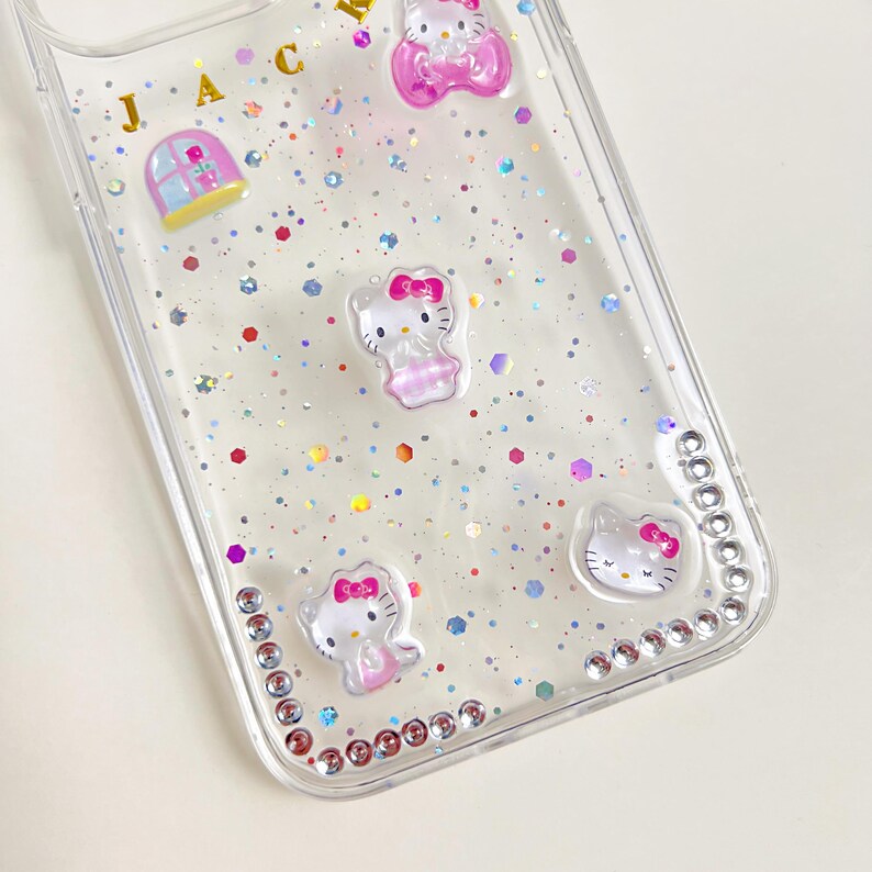 May include: Clear phone case decorated with Hello Kitty figures, pink bows, and glitter. The case has a border of small, clear rhinestones and gold letters spelling out "JAC".