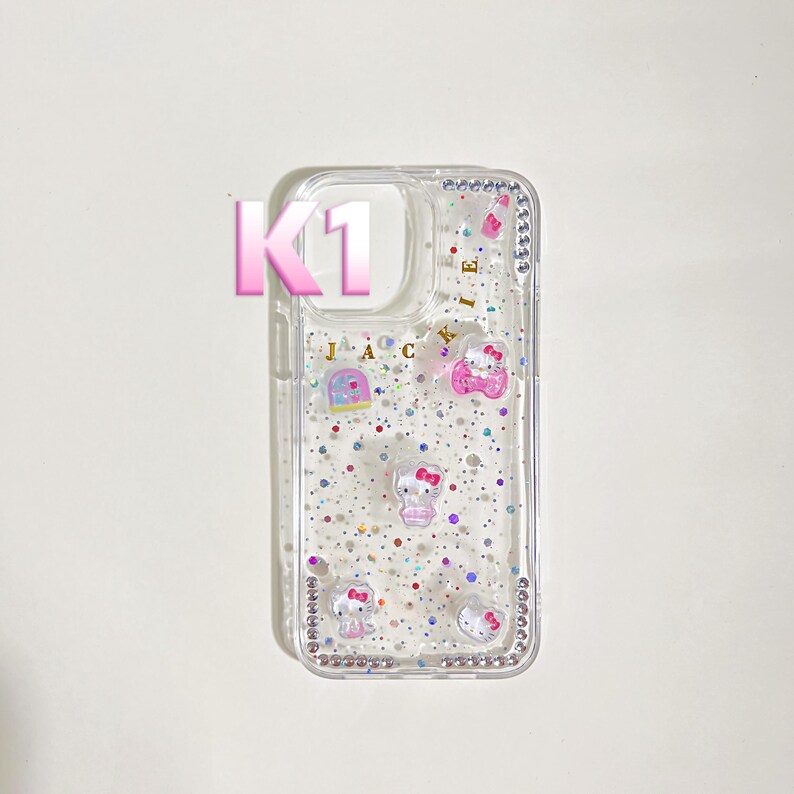 May include: Clear phone case with embedded glitter and Hello Kitty designs. The case features a rhinestone border and the text "J A C I E". The letter "K1" is also visible.