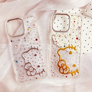May include: Two clear phone cases with a glittery background and a gold Hello Kitty design. The cases have a black and white polka dot pattern on the back.