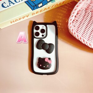 Puede incluir: Carcasa de tel&eacute;fono negra con fondo blanco, un lazo negro, un personaje Hello Kitty negro con un lazo rosa y una lente de c&aacute;mara con tres c&iacute;rculos negros.