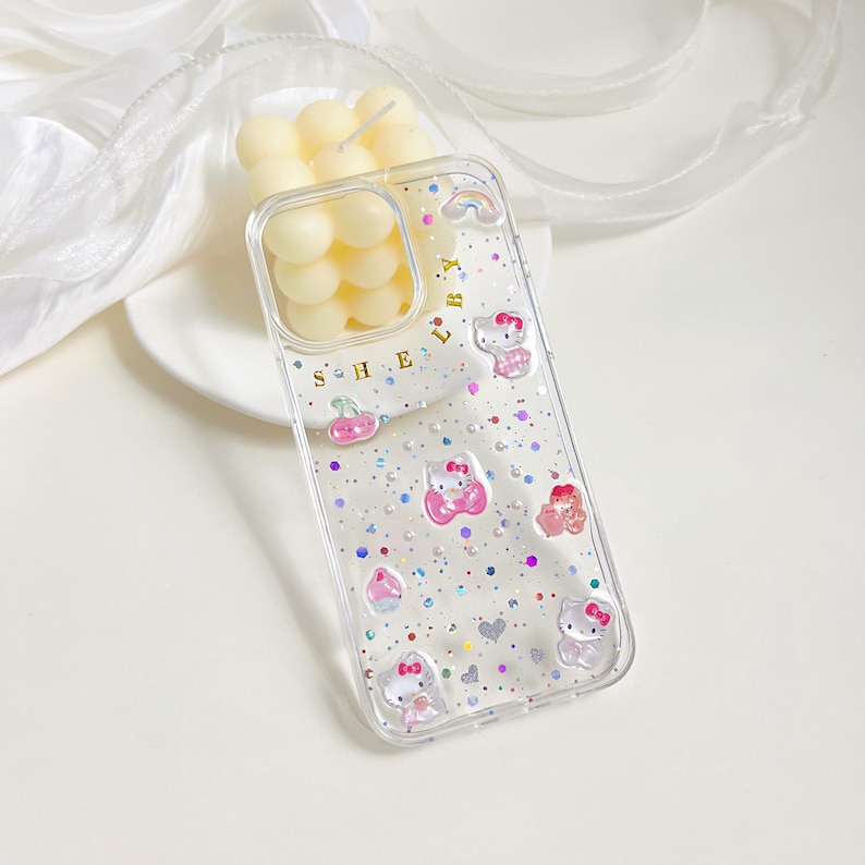 May include: Clear phone case decorated with Hello Kitty characters, pink hearts, and colorful glitter. The case has the letters "S H E L B Y" printed on it. The case is shown with a white plate and a candle.