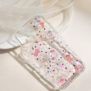 Peut inclure: Coque de t&eacute;l&eacute;phone transparente avec un fond scintillant rose et violet. La coque est orn&eacute;e de plusieurs petits charms, dont des c&oelig;urs, des &eacute;toiles et des personnages de dessins anim&eacute;s. Le texte "JESS" est &eacute;crit en lettres dor&eacute;es sur la coque.