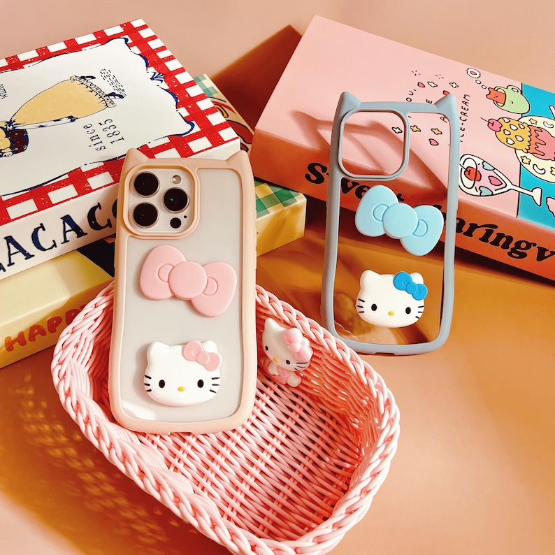 Puede incluir: Dos fundas transparentes para tel&eacute;fono con dise&ntilde;o de orejas de gato. Una funda es gris claro con un lazo rosa y un personaje de Hello Kitty. La otra funda es azul claro con un lazo azul y un personaje de Hello Kitty. Ambas fundas est&aacute;n en una cesta de mimbre rosa.