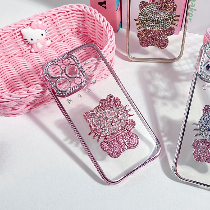 May include: Clear phone case with a pink frame and a sparkling Hello Kitty design. The case features a bedazzled Hello Kitty figure and a ring of rhinestones around the camera. A pink basket and Hello Kitty figurine are in the background.