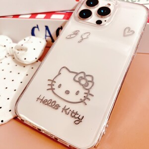 May include: Clear phone case with a silver Hello Kitty design. The case features a silver outline of Hello Kitty, a heart, and the text "Hello Kitty". The case is designed to fit a smartphone.