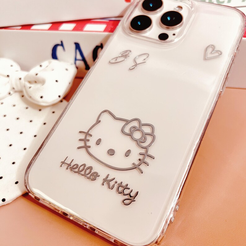 May include: Clear phone case with a silver Hello Kitty design. The case features a silver outline of Hello Kitty, a heart, and the text "Hello Kitty". The case is designed to fit a smartphone.