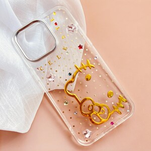 May include: A clear phone case with a gold Hello Kitty design and gold glitter. The case has a cutout for the camera lens and a gold inscription that says "Hello Kitty".
