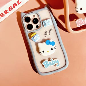 Op de afbeelding: Een lichtblauwe telefoonhoes met een wit Hello Kitty-personage, een blauwe strik en het woord "Baby" in blauwe letters. De hoes heeft ook een klein wit tasje met de letter "M" erop en een klein wit tasje met het woord "Sweet" erop. De hoes heeft een uitsparing voor de camera en een uitsparing voor de knoppen van de telefoon.
