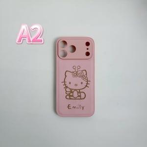 Puede incluir: Una funda de tel&eacute;fono rosa con un dise&ntilde;o de Hello Kitty y el nombre "Emily" grabado en dorado. La funda tiene recortes para la c&aacute;mara y los botones. El fondo es gris claro.