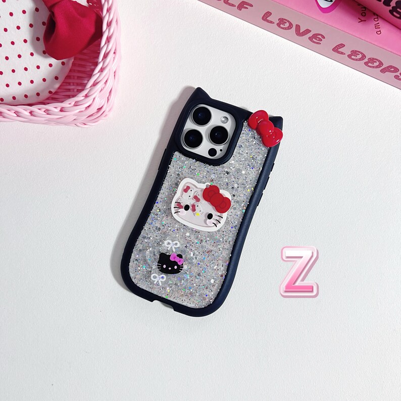 Może przedstawiać: Granatowe etui na telefon z błyszczącym srebrnym wnętrzem, z motywem Hello Kitty z czerwoną kokardą. Etui ma u g&oacute;ry kształt uszu kota i małą czerwoną kokardę z boku. Widoczna jest r&oacute;wnież r&oacute;żowa litera "Z".