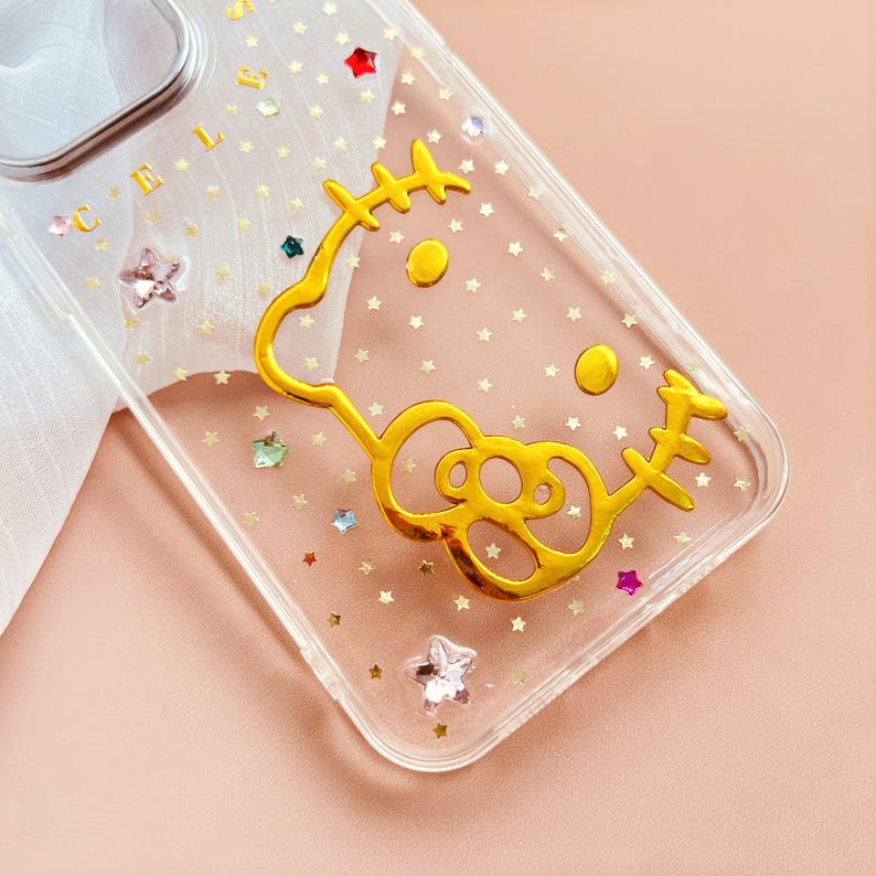 Pu&ograve; includere: Custodia per telefono trasparente con un design Hello Kitty dorato e stelle glitterate. La custodia ha la scritta "CELSI" stampata in alto.