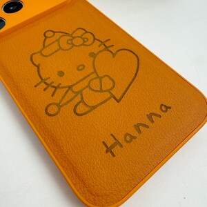 May include: Orange phone case featuring a cartoon character holding a heart, with the name "Hanna" engraved below. The design is in a darker shade of orange, creating a contrast against the case's background. The case has a smooth texture.