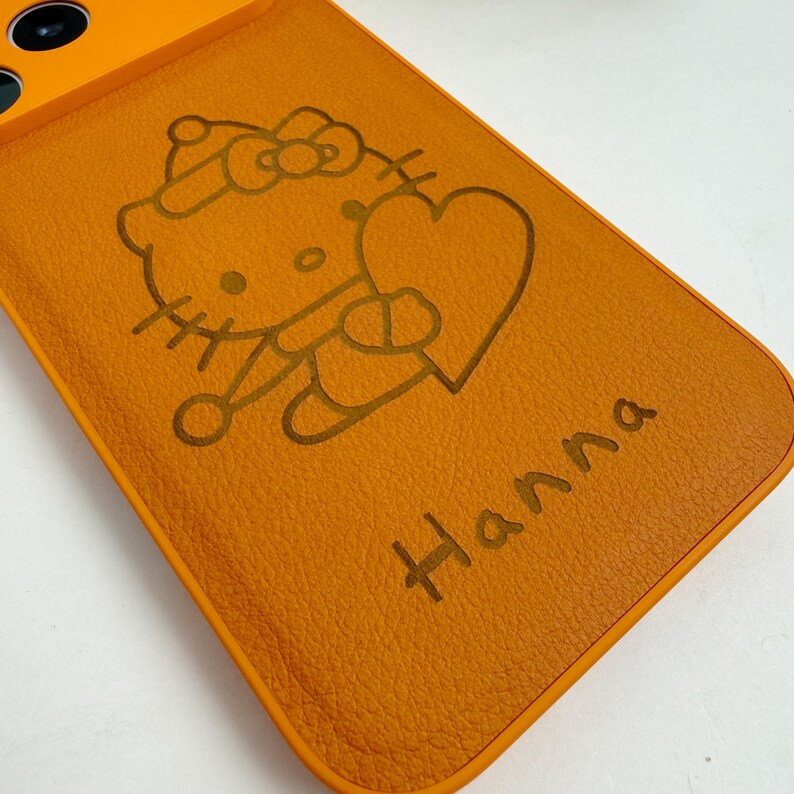 May include: Orange phone case featuring a cartoon character holding a heart, with the name "Hanna" engraved below. The design is in a darker shade of orange, creating a contrast against the case's background. The case has a smooth texture.