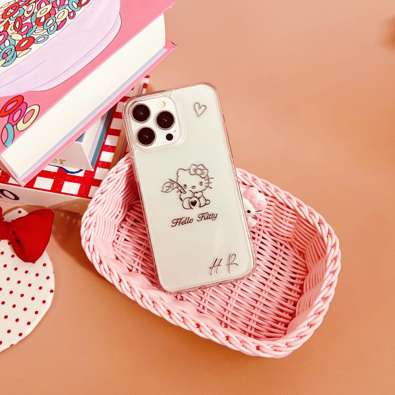 Op de afbeelding: Een transparante telefoonhoes met een Hello Kitty-ontwerp. De hoes heeft een roze hartje en de woorden "Hello Kitty" erop gedrukt. De hoes ligt in een roze rieten mand.