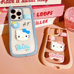 Op de afbeelding: Twee telefoonhoesjes met Hello Kitty-ontwerpen. Het blauwe hoesje heeft een witte Hello Kitty met een blauwe strik en de tekst "Baby". Het roze hoesje heeft een witte Hello Kitty met een roze strik en de tekst "Baby".