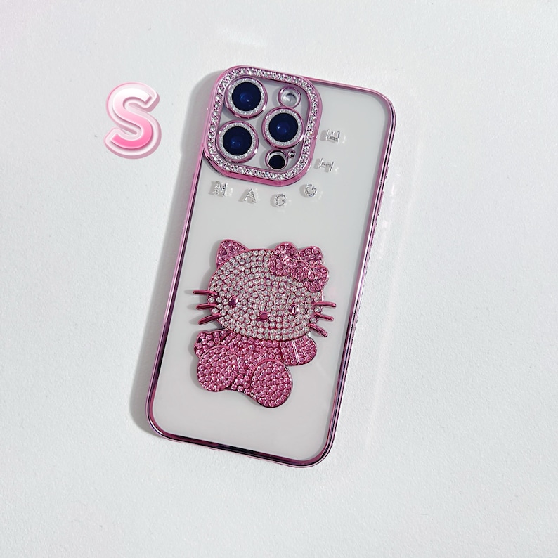 Pu&ograve; includere: Una custodia per telefono bianca con un design Hello Kitty rosa e una copertura per l'obiettivo della fotocamera in strass rosa. La custodia ha la scritta "MAG" stampata.