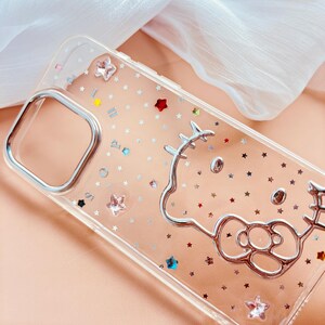 May include: Clear phone case with a silver Hello Kitty outline and silver glitter stars. The case has a silver trim around the camera lens and a silver trim around the edges.