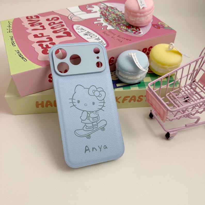 Pu&ograve; includere: Custodia per telefono azzurra con un design di Hello Kitty su uno skateboard e il nome "Anya". La custodia &egrave; accanto a oggetti decorativi, tra cui candele a forma di macaron e un piccolo carrello della spesa. La custodia ha un ritaglio per la fotocamera.