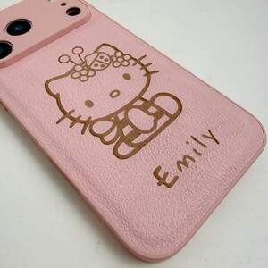 Puede incluir: Funda de tel&eacute;fono rosa con un contorno marr&oacute;n de un gato de dibujos animados con un lazo y el nombre "Emily" en fuente manuscrita. La funda tiene una superficie texturizada y un recorte para la c&aacute;mara.