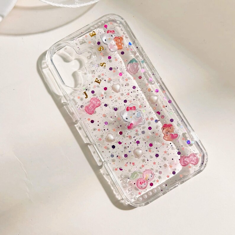 Peut inclure: Coque de t&eacute;l&eacute;phone transparente avec un fond scintillant rose et violet. La coque est orn&eacute;e de plusieurs petits charms, dont des c&oelig;urs, des &eacute;toiles et des personnages de dessins anim&eacute;s. Le texte "J.E.S." est imprim&eacute; en or sur la coque.