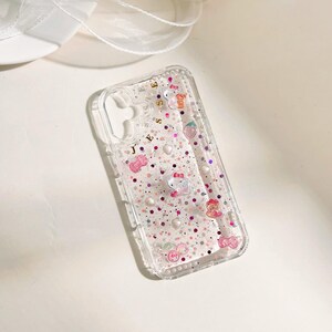 May include: A clear phone case with a pink, white, and gold glitter design. The case has a variety of small charms, including a strawberry, a heart, and a bow. The text "JESS" is written in gold on the case.