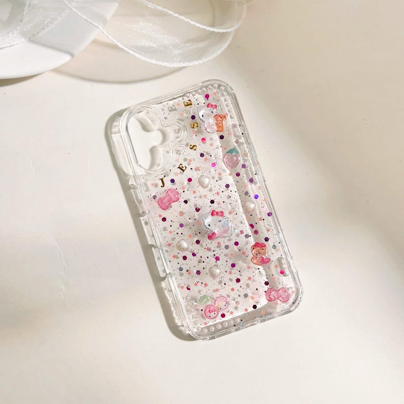 May include: A clear phone case with a pink, white, and gold glitter design. The case has a variety of small charms, including a strawberry, a heart, and a bow. The text "JESS" is written in gold on the case.