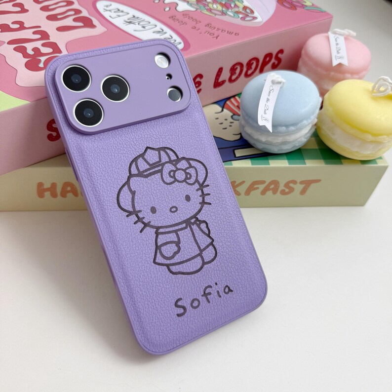 May include: A lavender phone case featuring a Hello Kitty graphic and the name "Sofia" in black. The case has a textured finish and a camera module. The background includes decorative macarons and a cereal box.