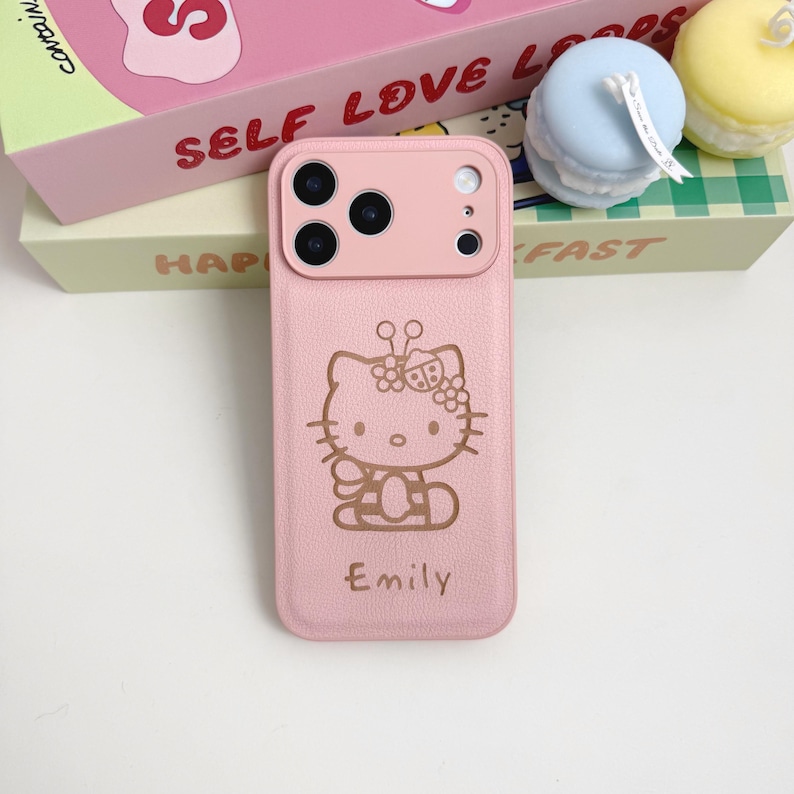 Puede incluir: Funda de tel&eacute;fono rosa con un dise&ntilde;o de Hello Kitty y el nombre "Emily" impreso debajo. La funda tiene un recorte para la c&aacute;mara. Otros art&iacute;culos visibles incluyen una caja rosa y un macarr&oacute;n azul.