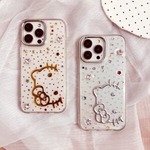 May include: Two clear phone cases with a Hello Kitty design. The cases are decorated with glitter, rhinestones, and stars. One case has gold accents and the text "CELESTE" on the back. The other case has silver accents and the text "SOPHIA" on the back.