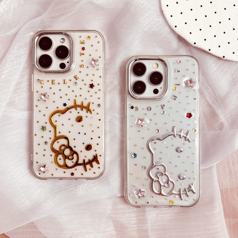 May include: Two clear phone cases with a Hello Kitty design. The cases are decorated with glitter, rhinestones, and stars. One case has gold accents and the text "CELESTE" on the back. The other case has silver accents and the text "SOPHIA" on the back.