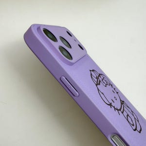 May include: A lavender phone case featuring a black outline illustration of a cartoon character. The case has a camera cutout and button details. The phone case is made of a smooth material.