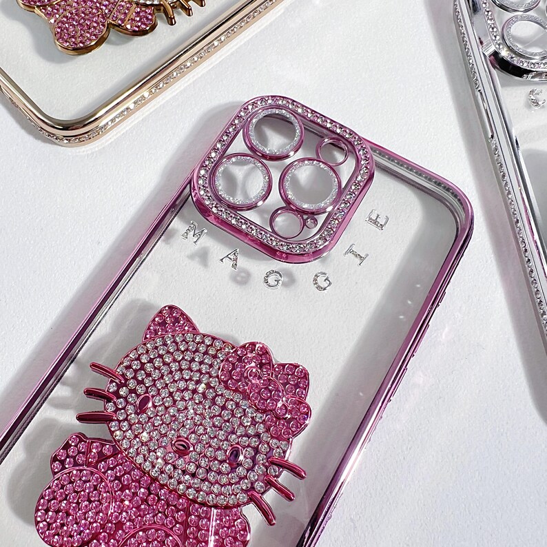 K&ouml;nnte beinhalten: Nahaufnahme mehrerer klarer Handyh&uuml;llen mit dekorativen Verzierungen. Eine H&uuml;lle zeigt ein pinkfarbenes Hello Kitty-Design, das mit Strasssteinen besetzt ist. Eine andere H&uuml;lle hat einen pinkfarbenen Kameraschutz mit Strasssteinen. Die H&uuml;llen haben eine transparente R&uuml;ckseite und einen metallischen Rand.