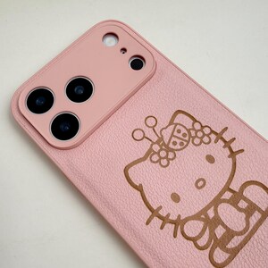 Puede incluir: Funda de tel&eacute;fono rosa con un dise&ntilde;o de Hello Kitty en relieve marr&oacute;n. La funda tiene una superficie texturizada y un m&oacute;dulo de c&aacute;mara con tres lentes. El dise&ntilde;o incluye una mariquita y flores.