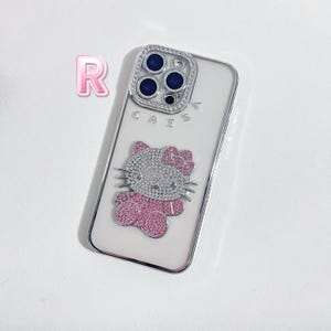 May include: A clear phone case with a silver frame and a Hello Kitty design. The Hello Kitty figure is pink and silver, with a bow. The camera lens is surrounded by a silver frame with rhinestones. The word "CAISA" is printed on the case.