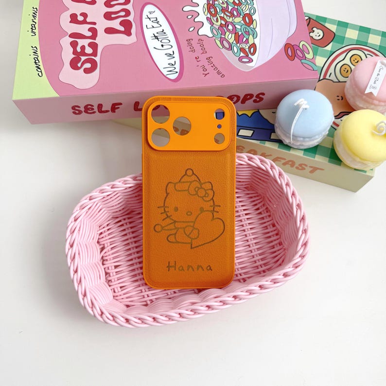 Puede incluir: Funda de tel&eacute;fono naranja con un dise&ntilde;o de Hello Kitty y el nombre "Hanna" en una cesta rosa tejida. La funda tiene un protector de lente de c&aacute;mara naranja. Una caja de cereales y macarons decorativos est&aacute;n en el fondo.