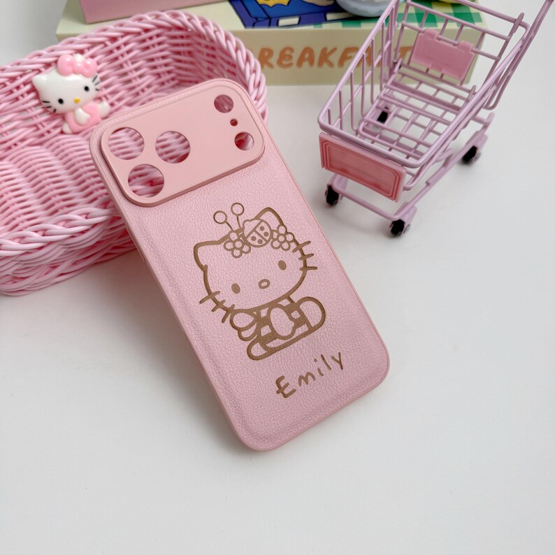 Puede incluir: Funda de tel&eacute;fono rosa claro con un dise&ntilde;o de Hello Kitty y el nombre "Emily" en dorado. La funda tiene recortes para la c&aacute;mara y est&aacute; rodeada de accesorios rosas, incluyendo un carrito de compras en miniatura y una cesta.